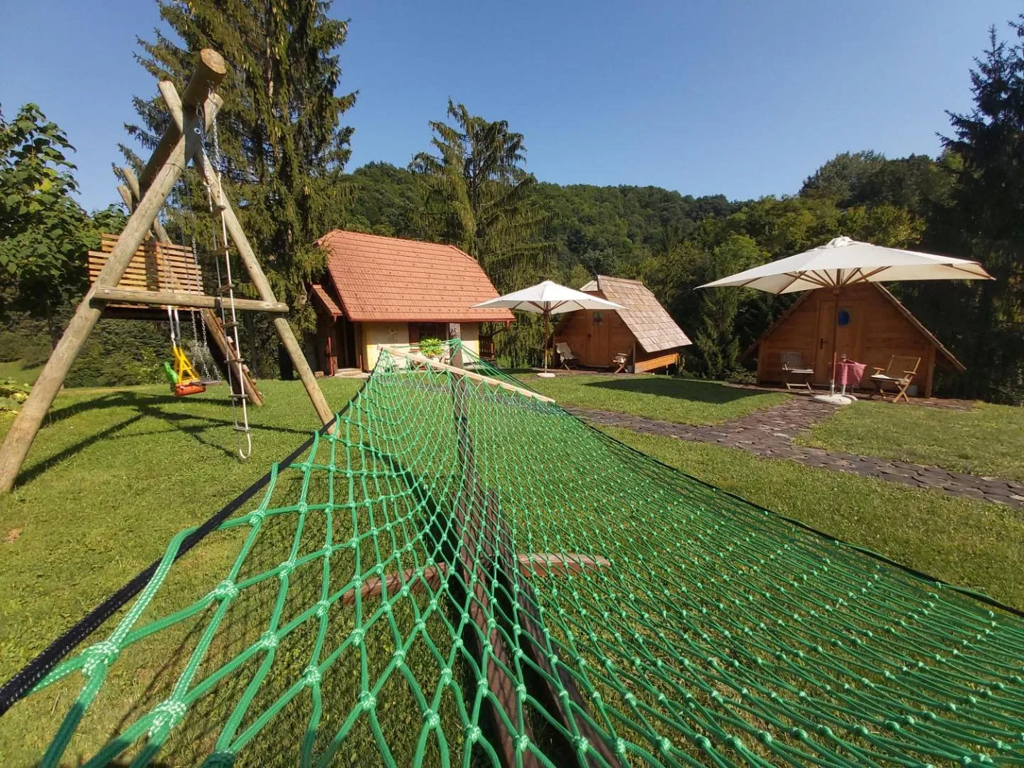Zelena mrežasta viseča mreža, napeta na travniku, za njo lesene glamping hiške, gugalnica in senčniki.