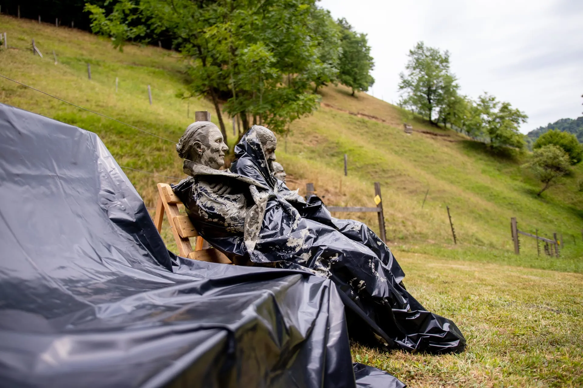 Woman covered in grey clay sitting in a small clay-bath pool, forest behind her.